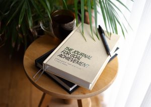 A sand-coloured “Journal for Goal Achievement” placed on top of a graphite journal on a small round table, accompanied by a coffee and a houseplant in the background.
