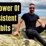 Andy Holt sitting on coastal rocks, wearing a black t-shirt and sunglasses, with a motivational quote overlay reading 'The Power of Consistent Habits'. The background features a sandy beach and a pier under a partly cloudy sky.