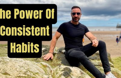 Andy Holt sitting on coastal rocks, wearing a black t-shirt and sunglasses, with a motivational quote overlay reading 'The Power of Consistent Habits'. The background features a sandy beach and a pier under a partly cloudy sky.